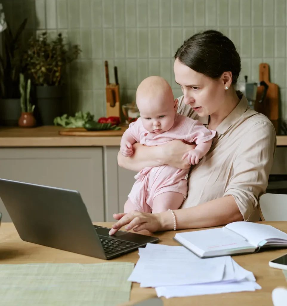 A mother holds her baby on one arm while she checks paperwork on a laptop at the kitchen table