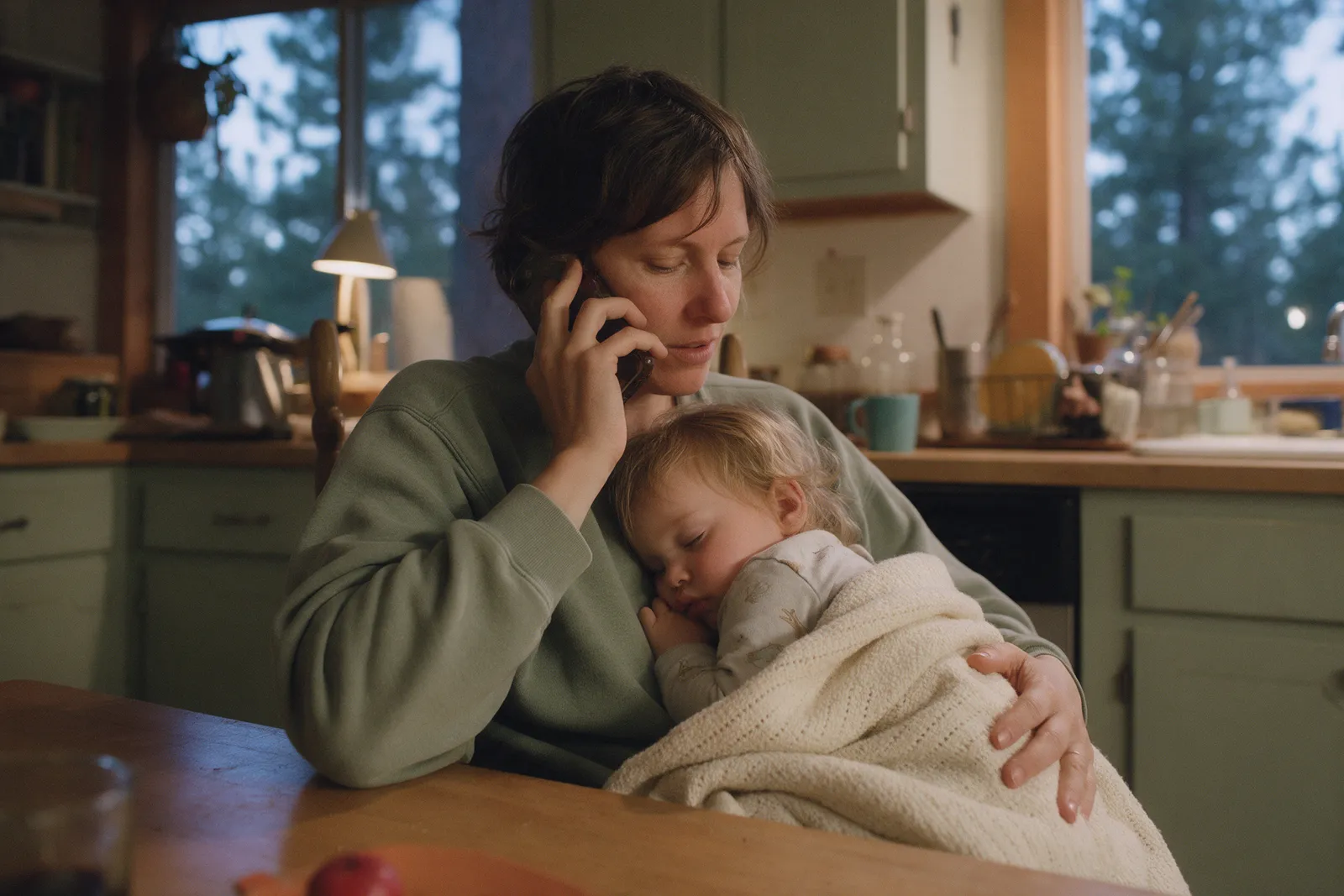 A parent on the phone at the kitchen table in the evening, holding a sleepy toddler wrapped in a blanket